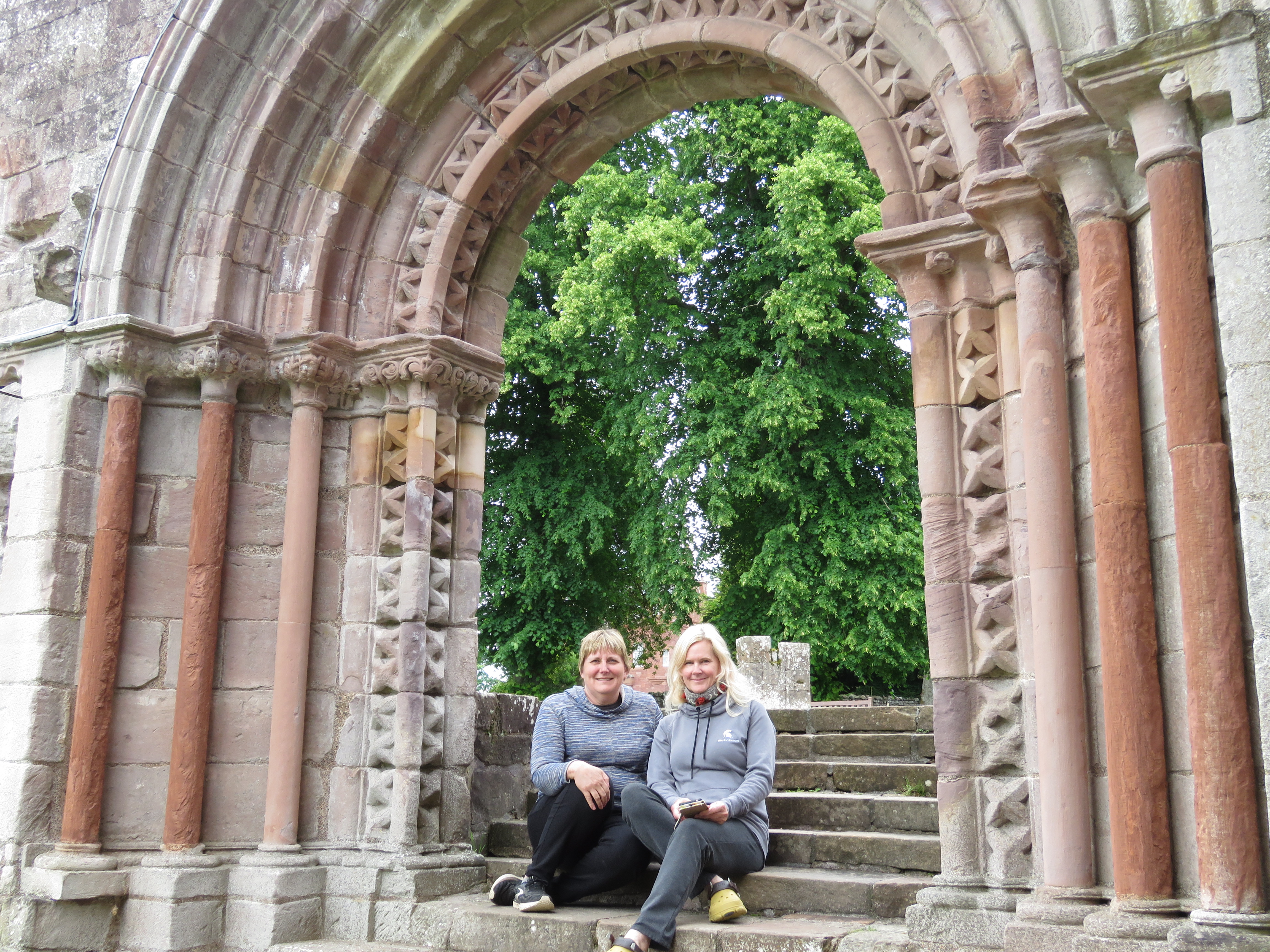 Two women sitting on stone steps beneath a large arched stone doorway with ornate carved columns. Behind them, green leafy trees fill the background.